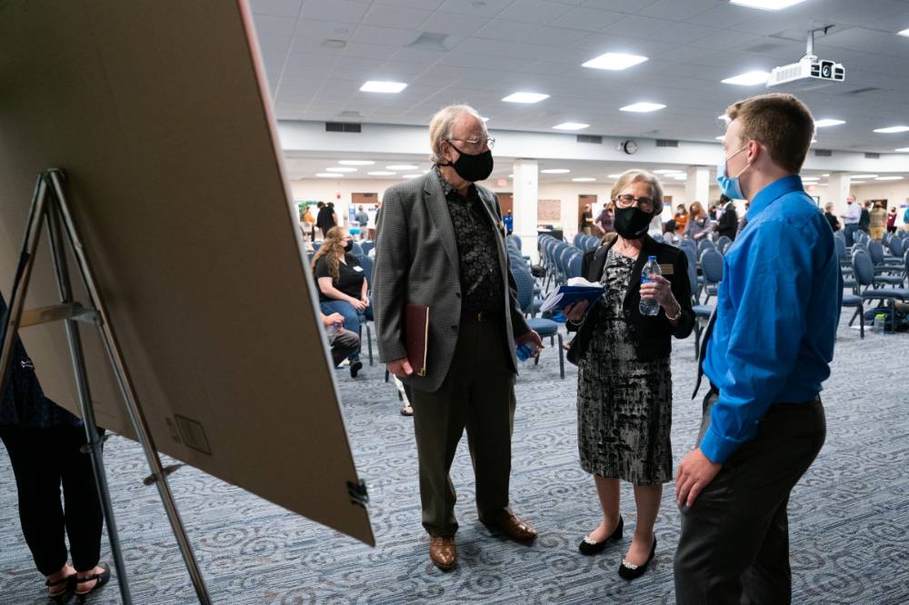 Scholar wearing mask presenting poster to two guests wearing masks.